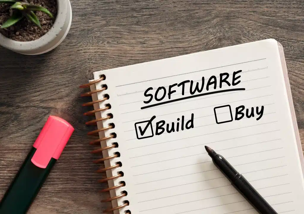 Notebook on a desk showing the word “Software” with a checked box next to “Build” and an unchecked box next to “Buy,” alongside a pen and highlighter.