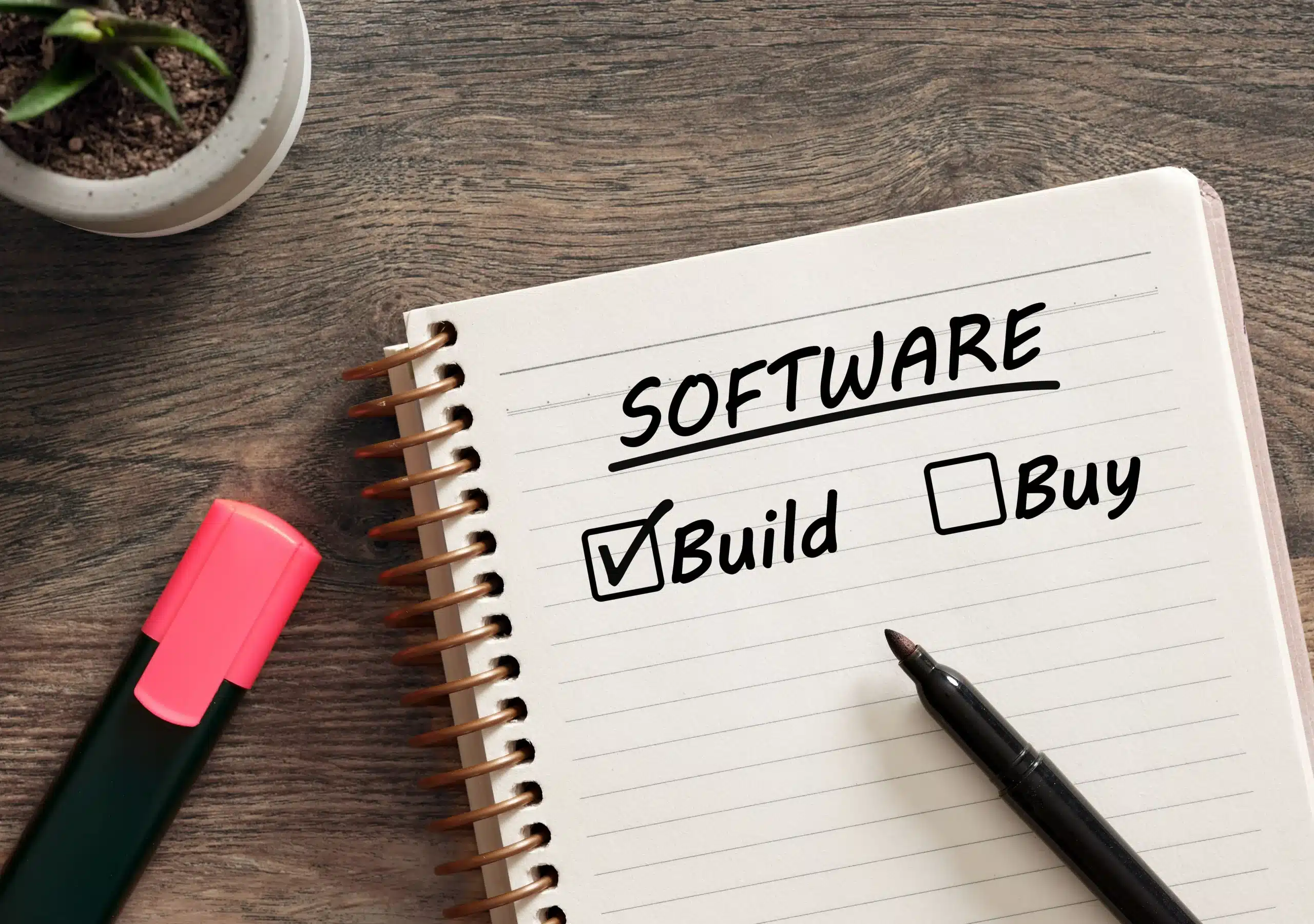 Notebook on a desk showing the word “Software” with a checked box next to “Build” and an unchecked box next to “Buy,” alongside a pen and highlighter.