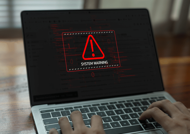 Laptop screen displaying a red “System Warning” alert with a caution symbol while a user types on the keyboard.